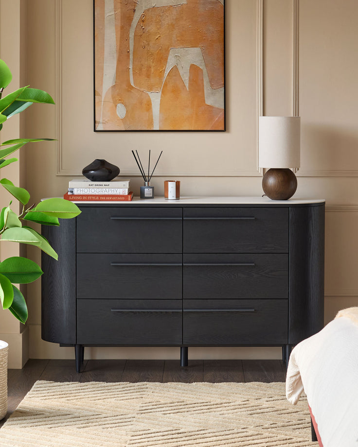 Black wood 6-drawer chest with ceramic white top, rounded sides, and tapered legs in a styled bedroom setting.