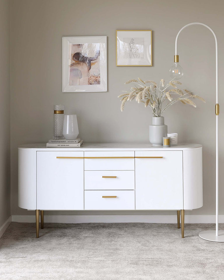 Elegant white sideboard with a curved silhouette featuring brass-finished legs and handle accents, enhanced by a tasteful arrangement of framed artwork, decorative books, vases, and an arching floor lamp.