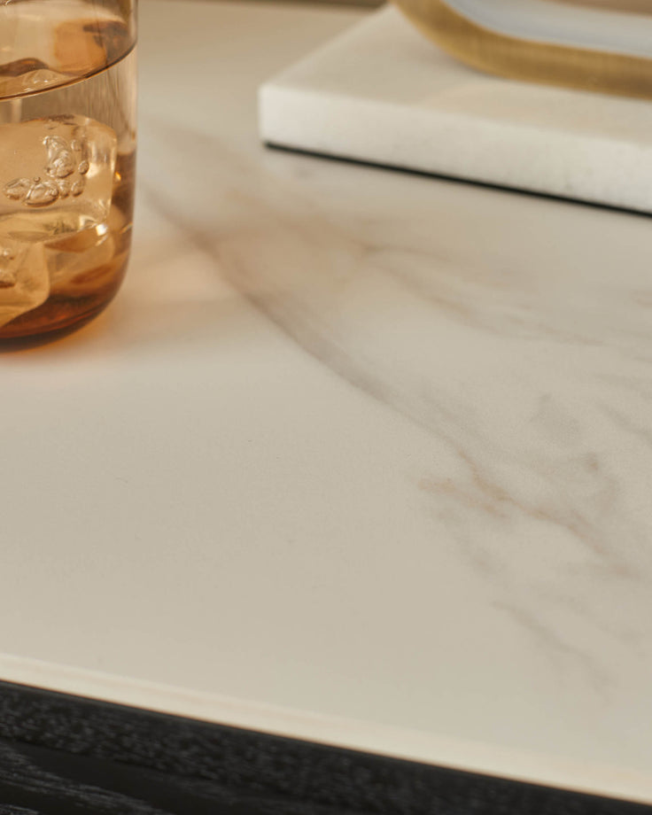 White marble bedside table with black base, styled with a glass of amber liquid and white napkin.