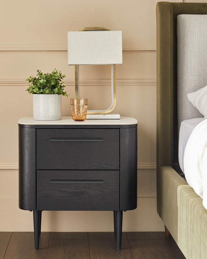 Black wooden bedside table with white ceramic top, two drawers, curved sides, and tapered legs in a bedroom setting.