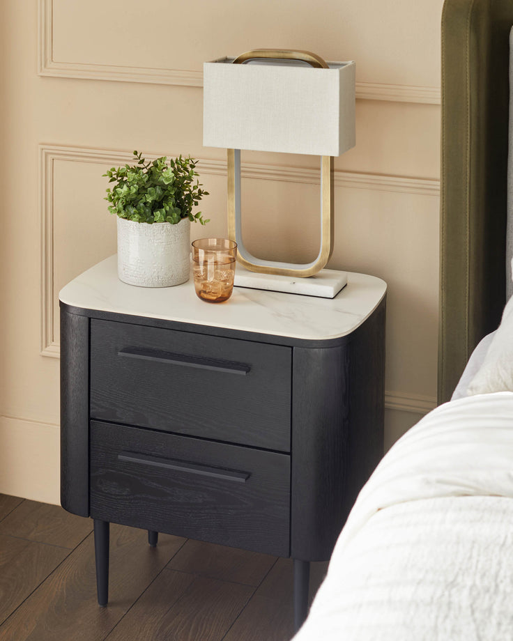 Black wood bedside table with white ceramic top, two drawers, and tapered legs, styled in a bedroom setting.