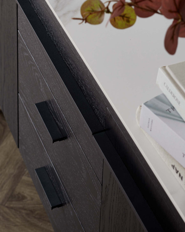Close-up view of a modern grey wooden cabinet with sleek black handles, featuring clean lines and a contemporary textured finish, partially displaying a marble surface on top with books and decorative foliage.