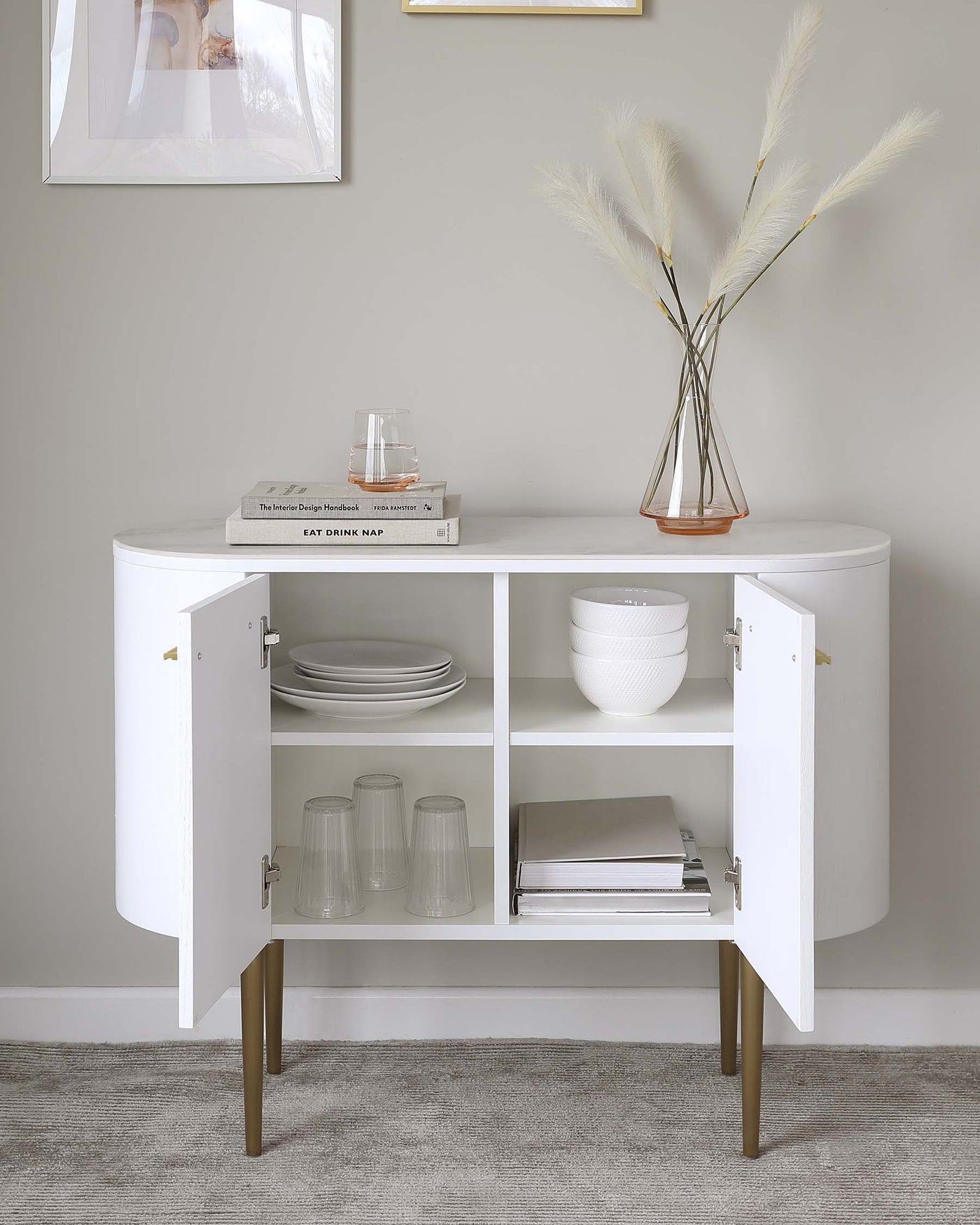 Elegant modern white sideboard with curved edges and half-circle cabinet doors, standing on splayed wooden legs. The sideboard is styled with neatly stacked books, glassware, and white ceramic dishes, complemented by a decorative glass vase with tall feather reeds.