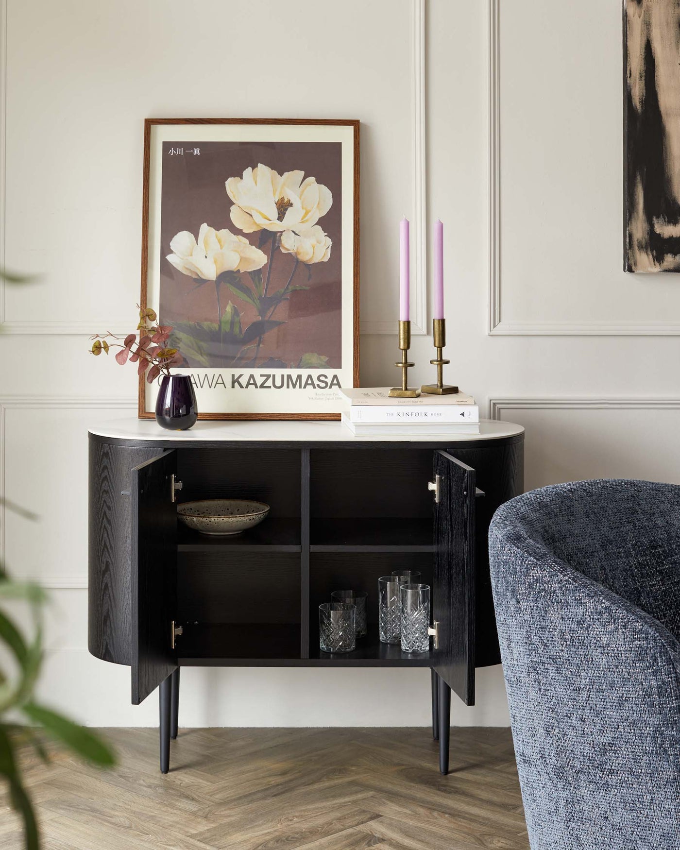 A sleek, semi-circular dark wooden console table with a white tabletop, featuring two cabinet doors with brass handles and slim, tapered legs. Shelves inside the cabinet display decorative items, and the table surface is styled with books, a purple vase with flowers, and a pair of pink candles in brass holders. A large framed floral artwork rests against the wall above the console.