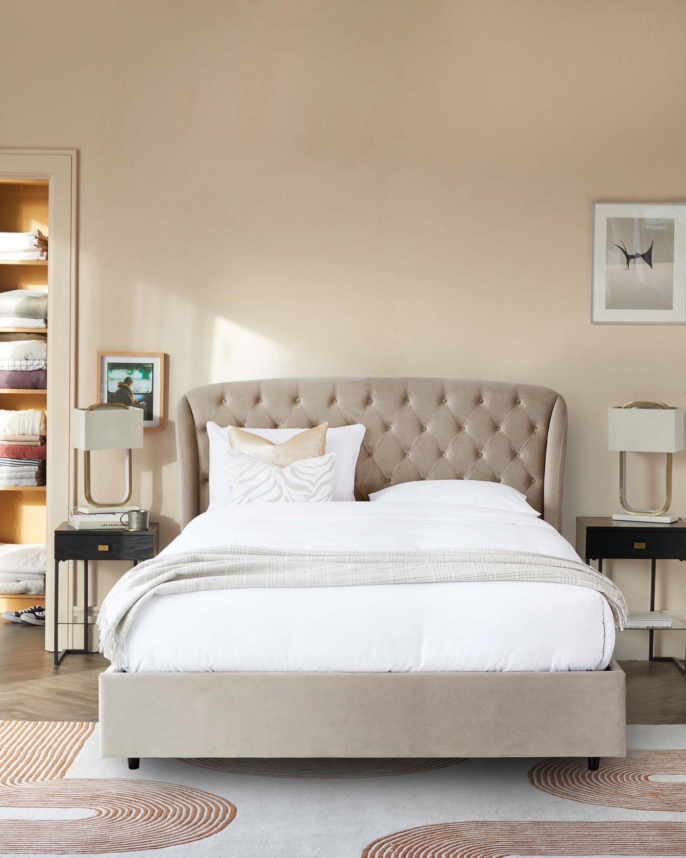 Elegant bedroom featuring a plush, tufted headboard on a large, neutral-toned upholstered bed. Coordinating nightstands with sleek, black frames and clear glass lamps flank the bed, creating a symmetrical, upscale look.