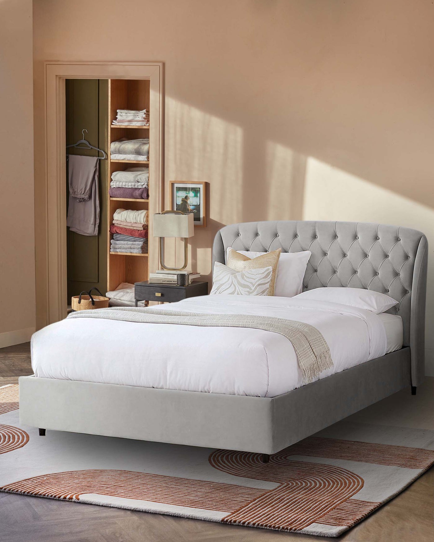 Elegant grey upholstered bed with a tufted headboard design and a neatly draped white bedsheet, complemented by decorative beige and golden throw pillows. There's a matching grey bedside table with a modern lamp and a subtle piece of art, placed on a contemporary rug with geometric patterns in shades of beige, orange, and grey.