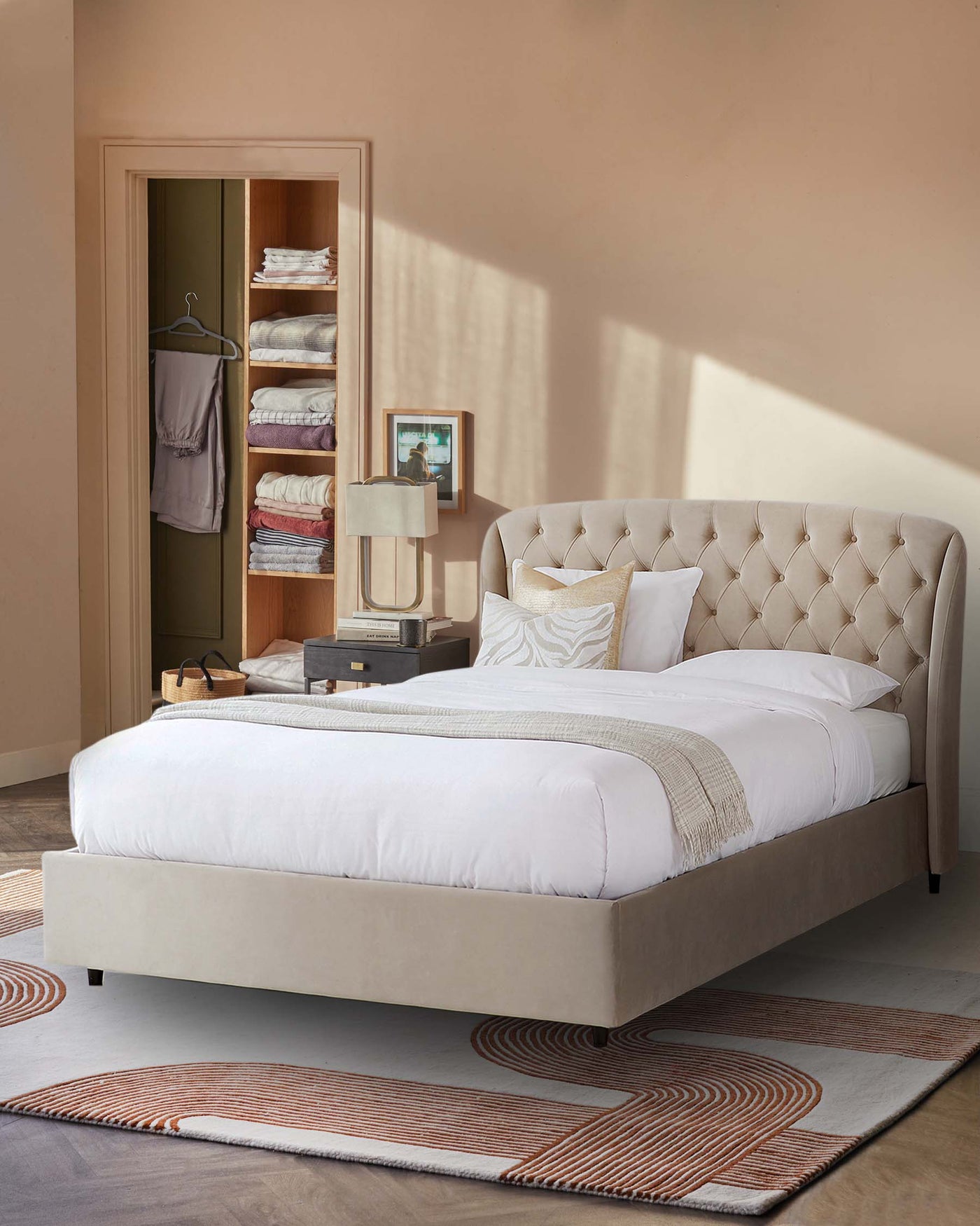 Elegant upholstered bed with tufted headboard in a neutral colour, featuring a low-profile design and clean lines. A stylish table lamp with a rectangular shade and metallic base rests on a stack of books next to the bed. The room is complemented by a contemporary area rug with a geometric pattern in warm tones.