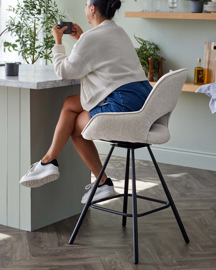A modern bar stool with textured light beige upholstery and a curved backrest. The stool features a sleek black metal frame with four tapered legs and built-in footrests, offering both support and contemporary style. It complements a marble-topped kitchen island in a cosy interior setting with wooden flooring.