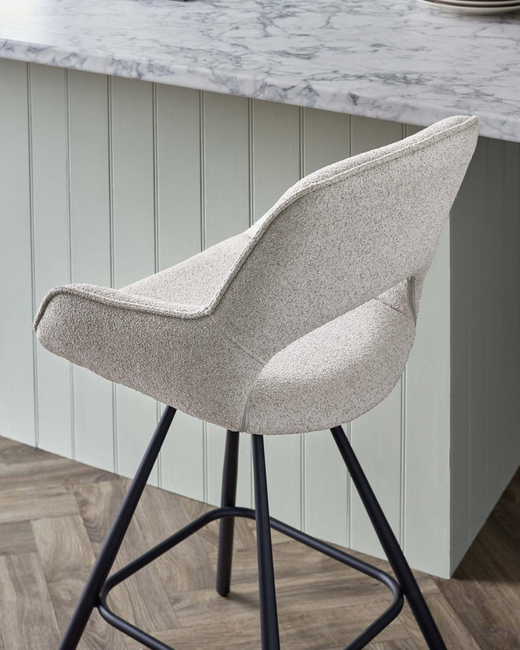 Modern upholstered bar stool with a textured light grey fabric and wingback design, featuring a supportive backrest and a well-cushioned seat. The stool stands on slender, black metal legs with a geometric base for stability. The contemporary style complements the marble countertop and pale green panelled bar backdrop.