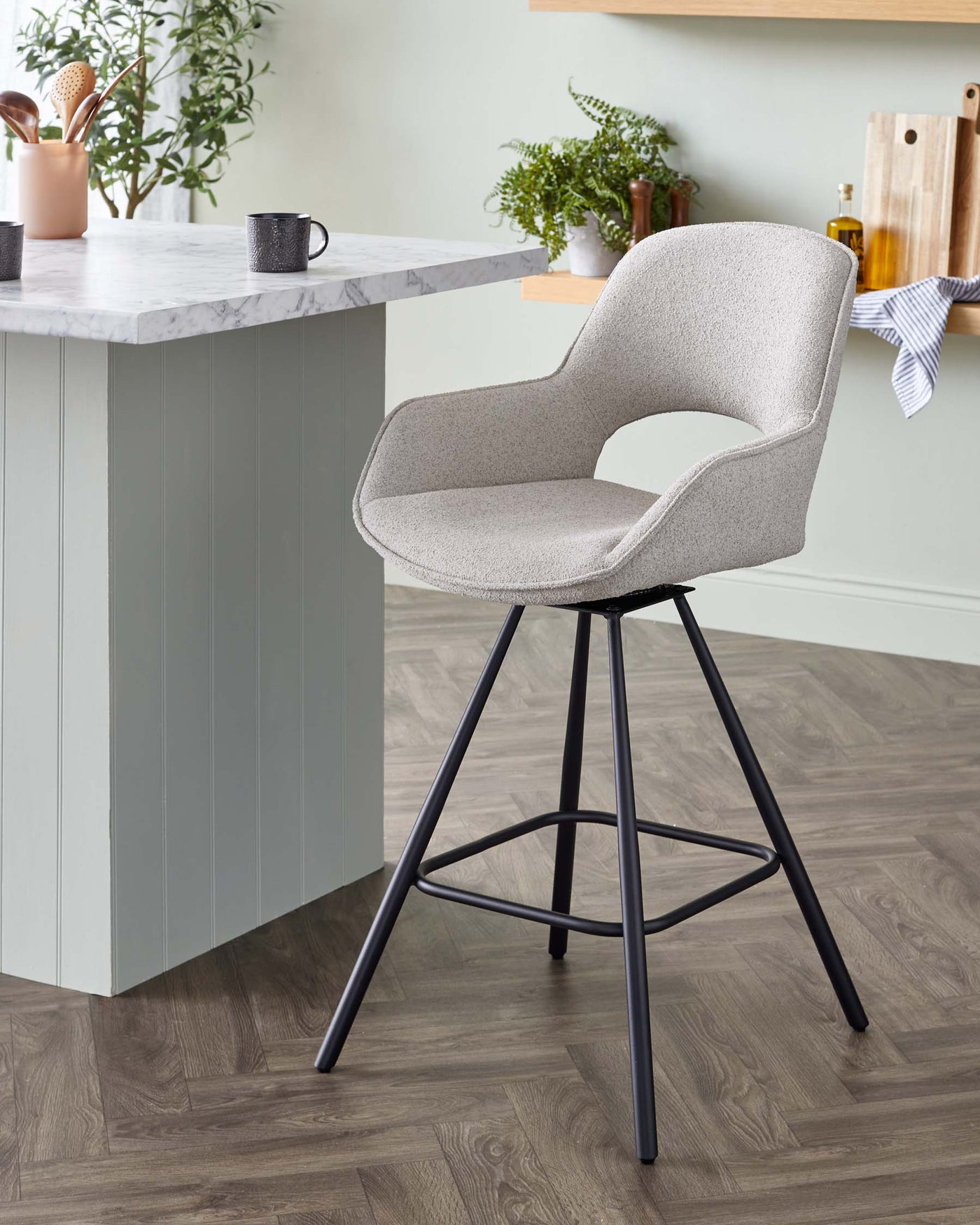 Modern bar stool with a textured light grey fabric upholstery and an ergonomic curved design. It features a swivel seat, armrests, and is supported by a slim, black metal frame with four slanted legs and a circular footrest. The chair is positioned by a kitchen counter with a marble top, showcasing a sophisticated and contemporary style suitable for kitchen or bar areas.