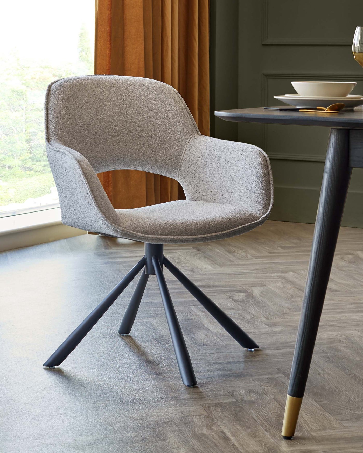 Modern upholstered dining chair with a curved backrest and a swivel base featuring four angled legs. The chair has a light grey textured fabric and matte black legs with contrasting metallic tips. Part of a round wooden dining table with a black finish is visible, set with a bowl and plate on top.