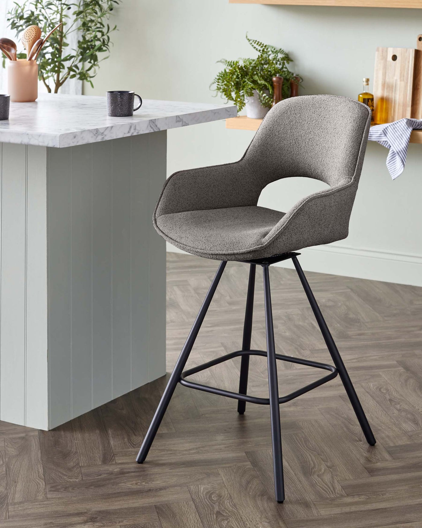 Modern upholstered bar stool with a curved backrest and armrests, featuring a speckled grey fabric. The stool has a slender black metal frame with four angled legs and a circular footrest. It is positioned next to a kitchen island with a marble countertop.