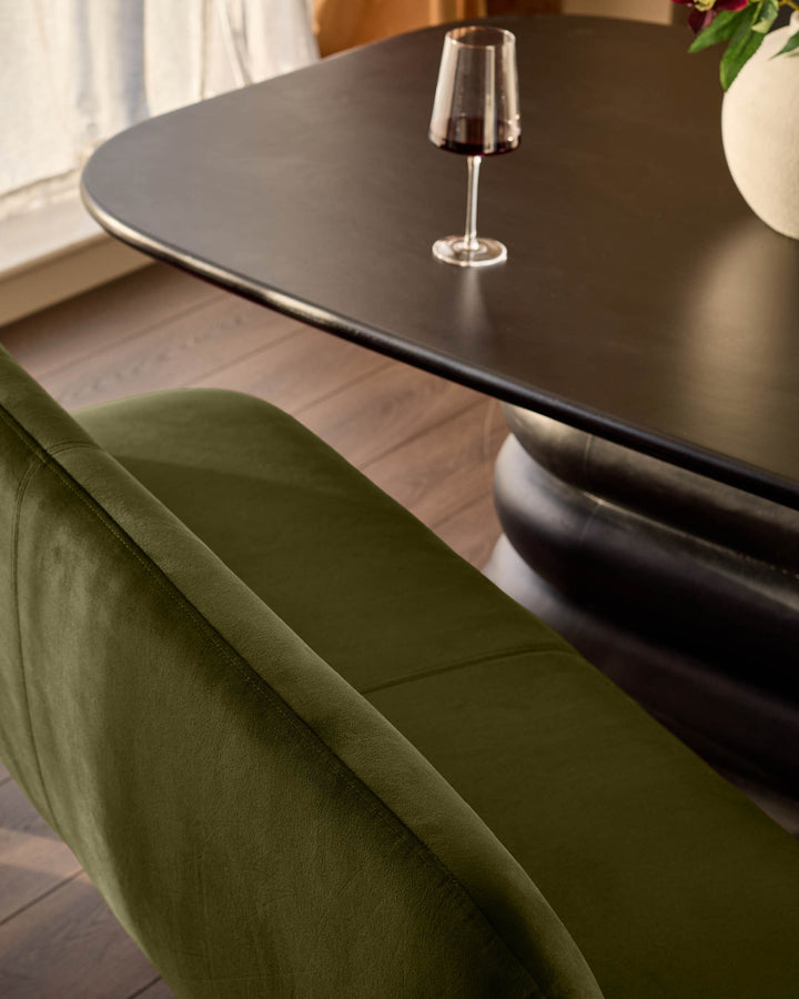 Mason 3-seater dining bench in olive green velvet, shown from side angle next to black table with wine glass.