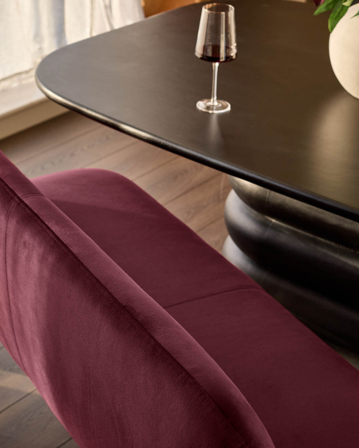 Mason 3-seater dining bench in plum velvet, shown from side angle next to black table with wine glass.