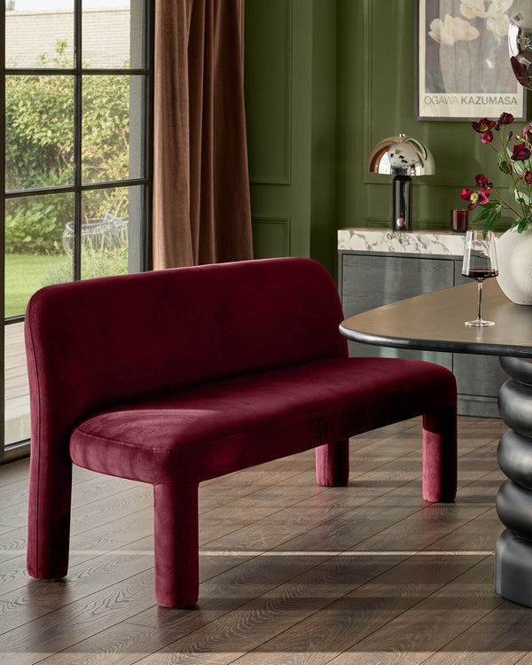 Mason 3-seater dining bench in plum velvet with curved backrest and cylindrical legs, positioned in green interior space.