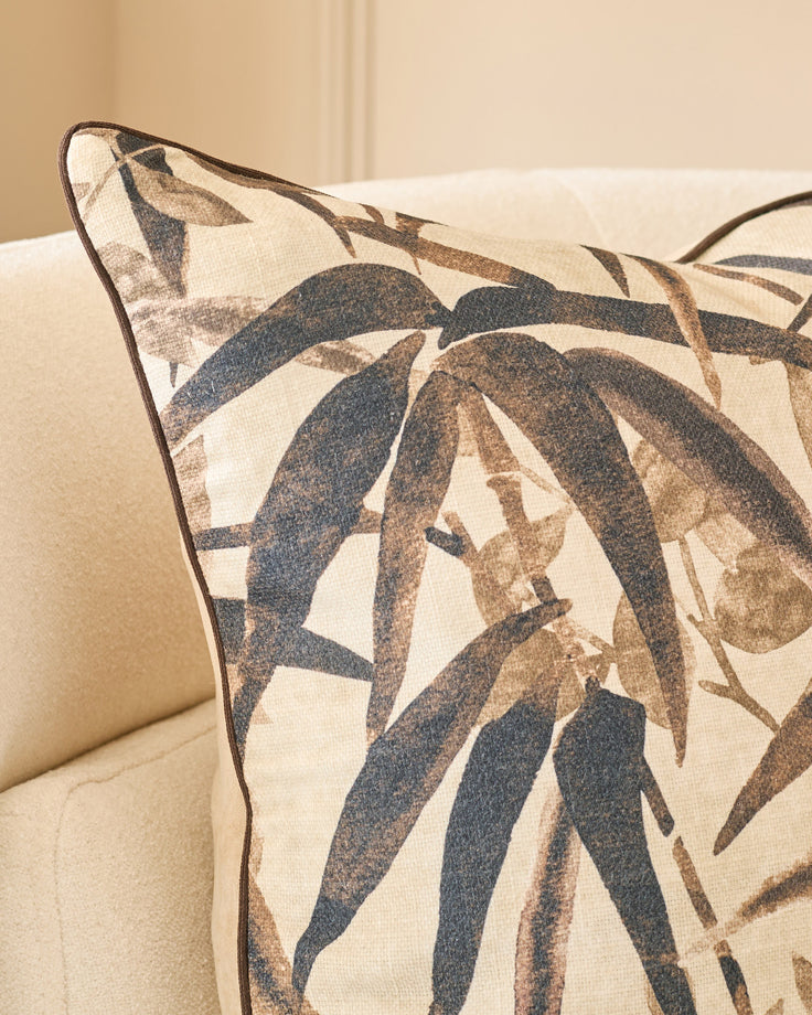 Madeira cushion in natural tones featuring dark blue and taupe leaf pattern on cream linen, displayed on beige upholstered furniture.