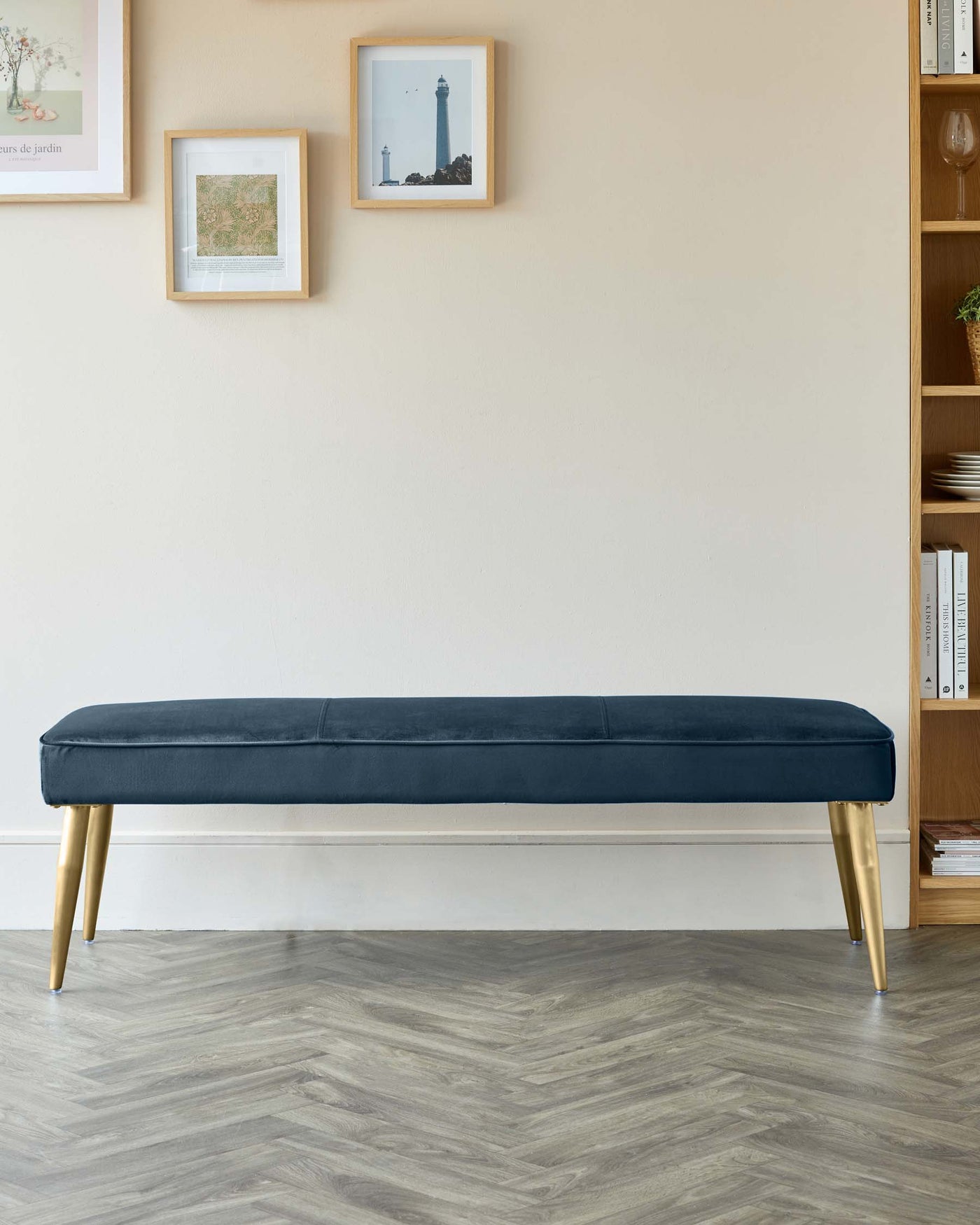 A contemporary navy blue upholstered bench with plush cushioning and sleek gold-tipped legs, positioned against a neutral wall with framed artwork and adjacent to a wooden bookshelf.