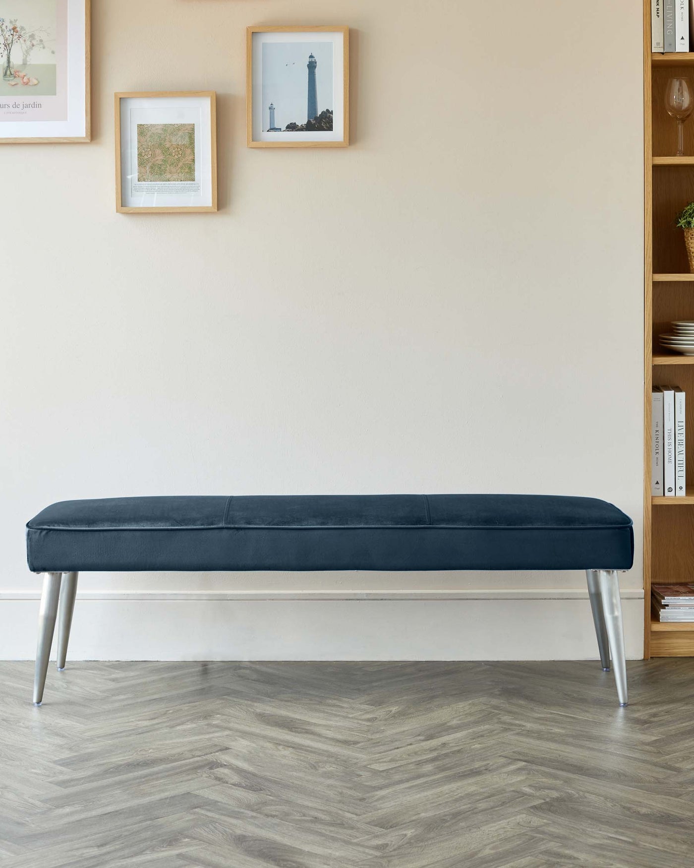 Modern minimalist upholstered bench in dark blue velvety fabric with sleek cylindrical metallic legs on a herringbone pattern wooden floor, complemented by a wooden bookshelf on the side.