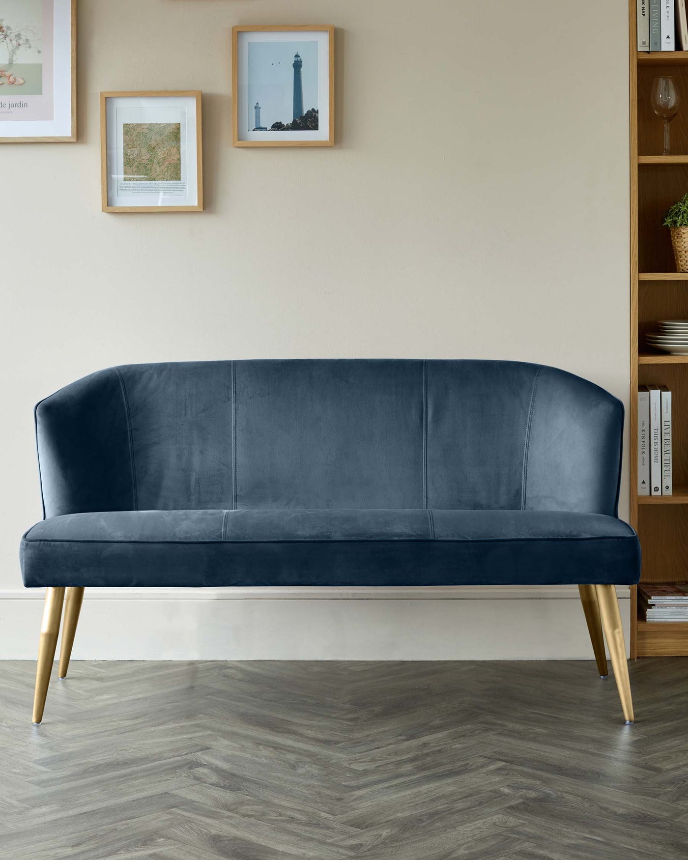 Elegant, mid-century inspired navy blue velvet sofa with a curved backrest, smooth seat, and angled gold metal legs, displayed in a contemporary living room setting.