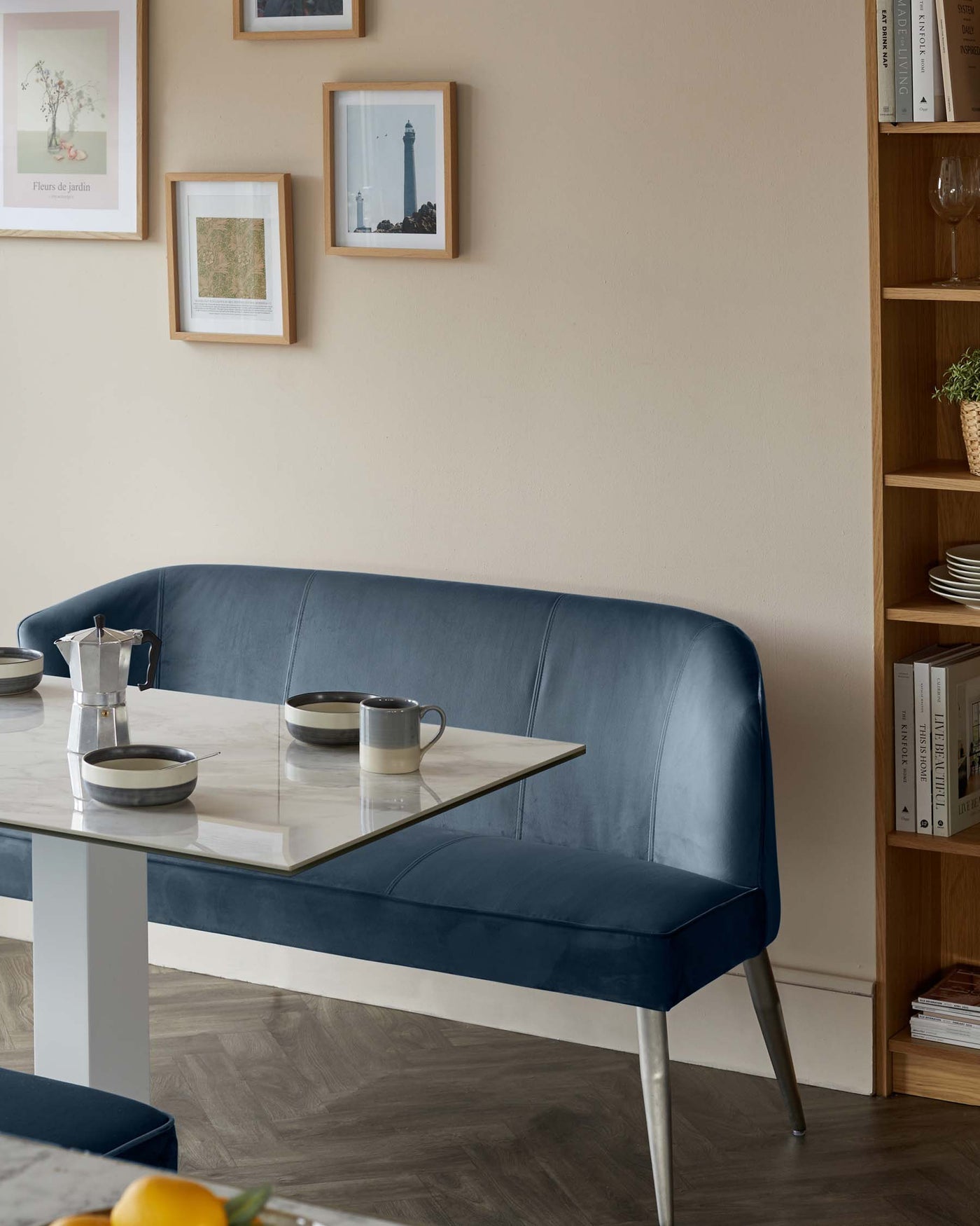 Modern dining area featuring a sleek white marble tabletop on a streamlined metal base, paired with a contemporary blue upholstered bench with slender metal legs. Adjacent to the dining set, a wooden open shelving unit with a variety of decorative items and books adds warmth to the space.