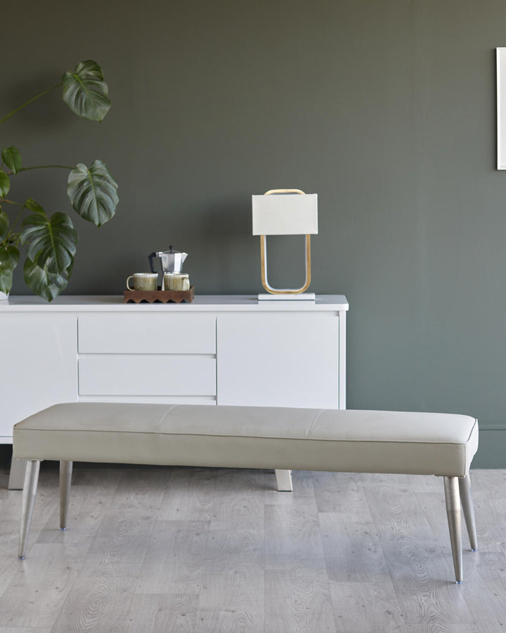 Contemporary white sideboard with decor, positioned behind a stylish grey bench with silver legs. Minimalist, elegant design.