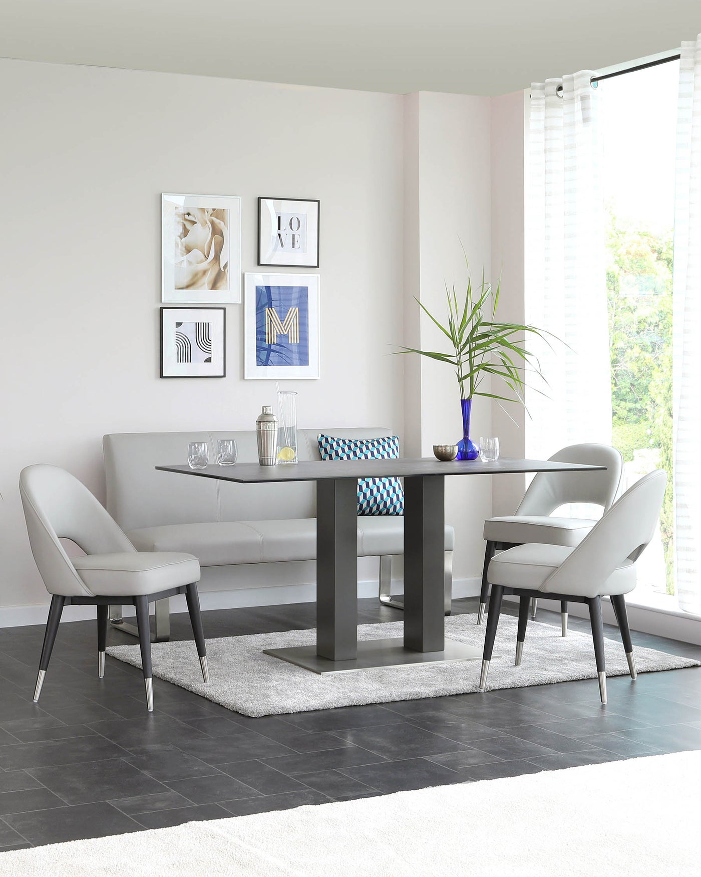 Modern dining room furniture featuring a sleek black table with a rectangular top and wide, flat legs, paired with three light grey upholstered chairs with curved backs and metallic legs. A matching light grey upholstered bench is set against the wall under framed artwork. The set is arranged on a plush grey area rug over dark tiled flooring.