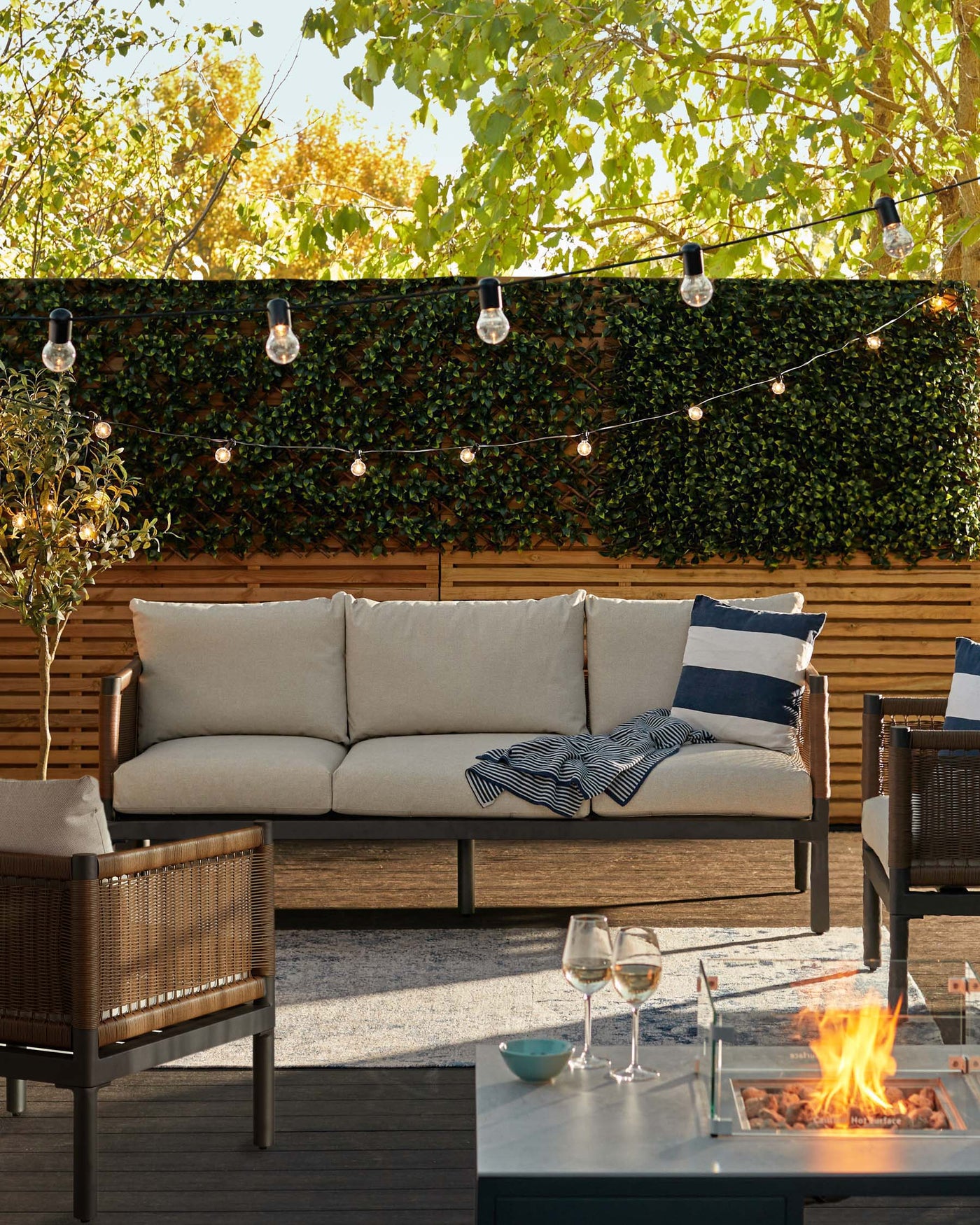 Contemporary outdoor furniture set featuring a large three-seater sofa with beige cushions and wooden armrests, a single armchair with matching design and cushions, and a rectangular coffee table with a dark surface and a built-in fire pit. The set is presented on a wooden deck with decorative string lights above, creating an inviting ambiance.