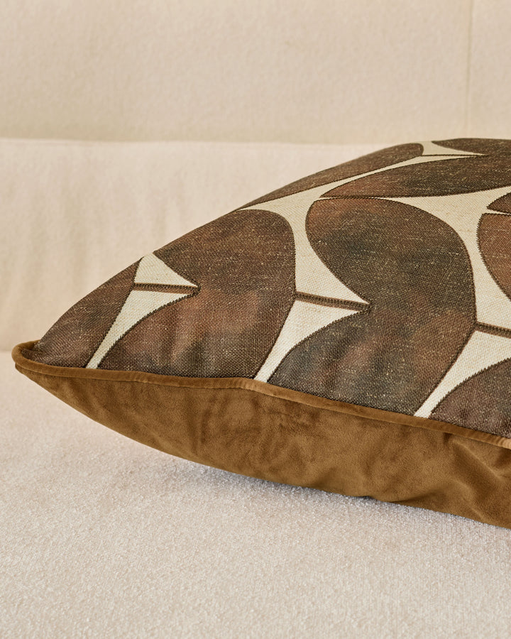 Micah cushion in chocolate featuring geometric leaf pattern with cream piping detail, shown at angle on light surface.