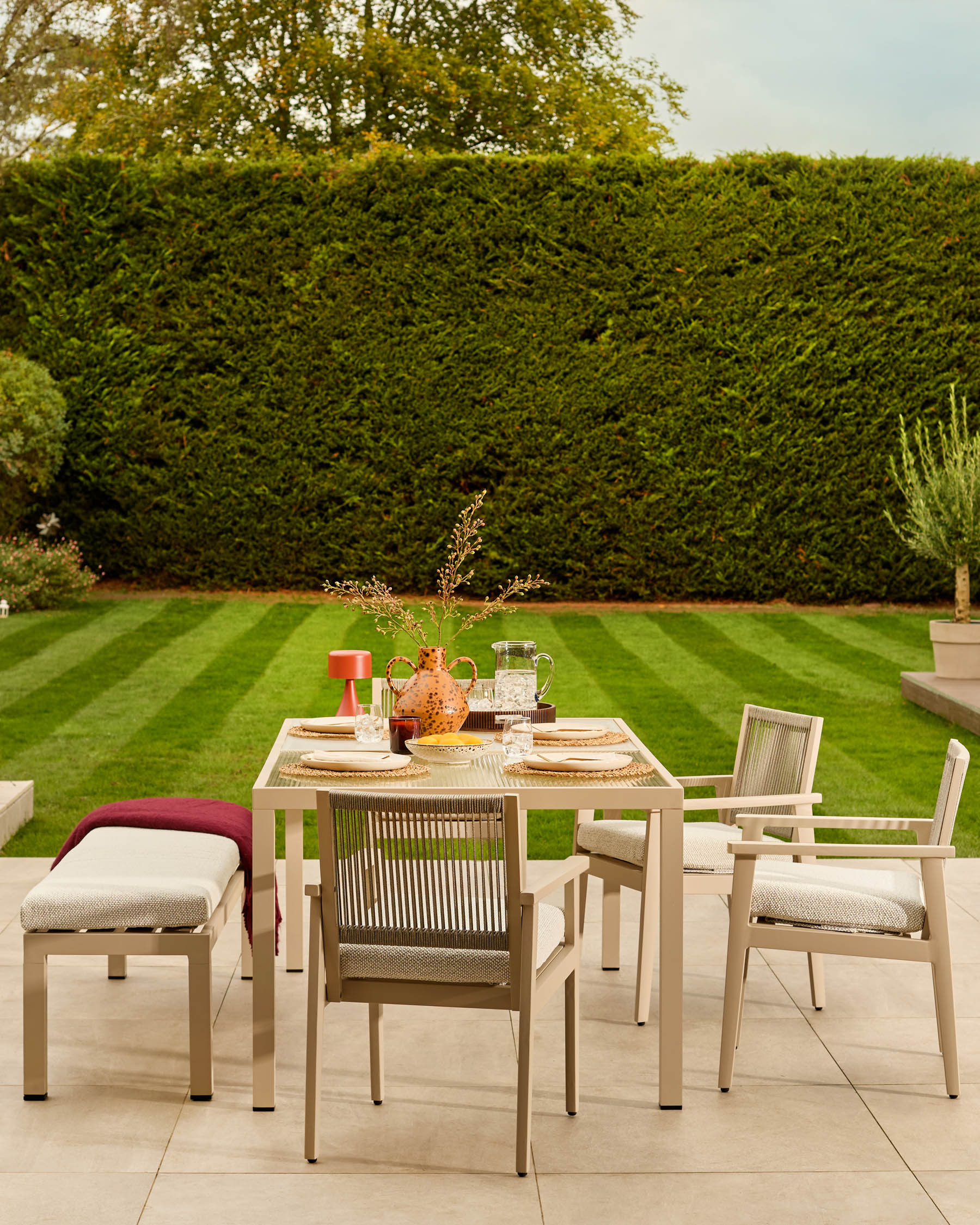 Minori natural garden dining bench with burgundy cushion, styled in an outdoor patio setting with matching chairs and dining table.
