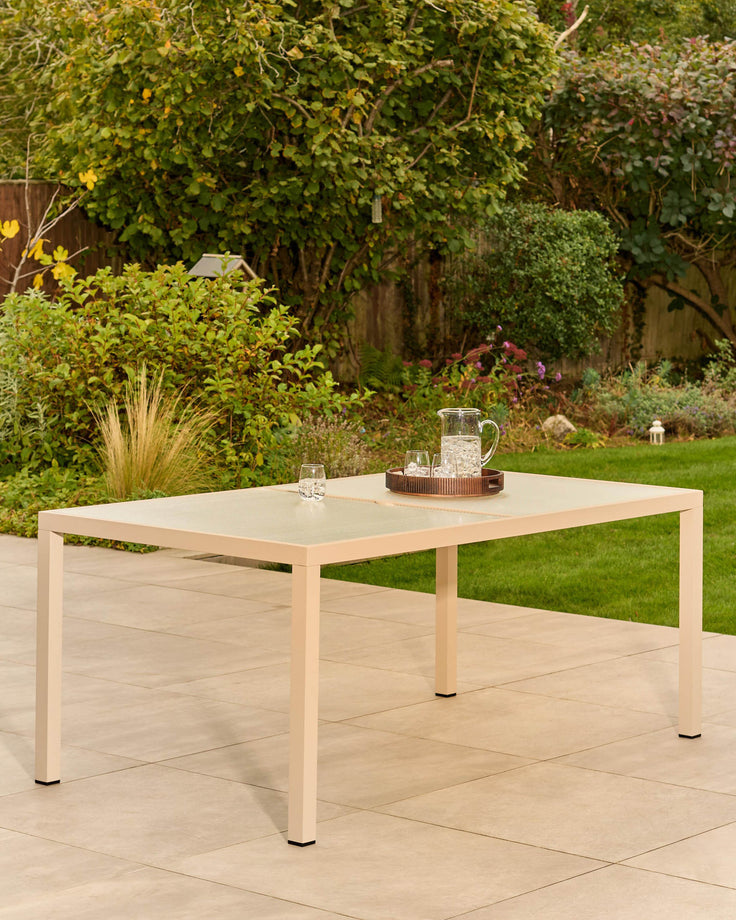 Cream-colored rectangular outdoor dining table with glass top and wooden frame, set on a paved patio with lush garden background.