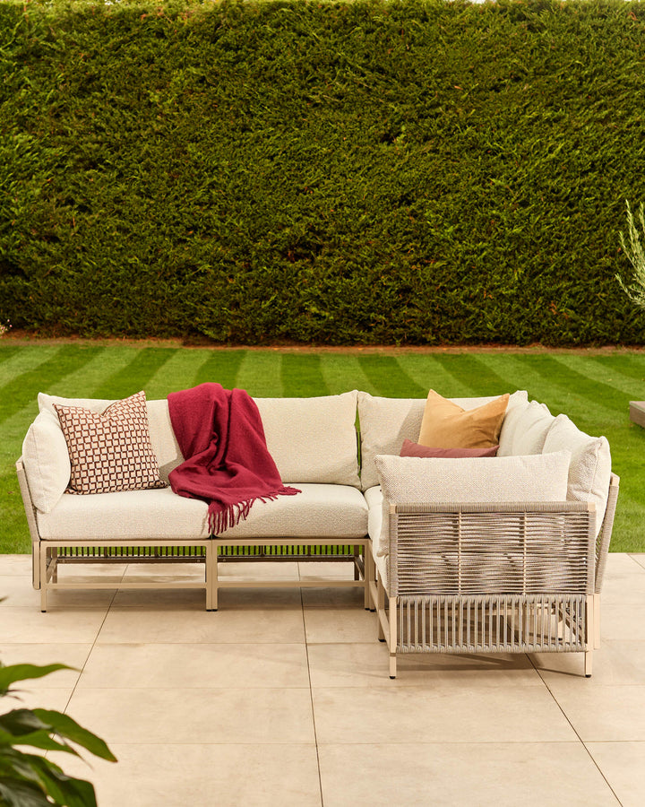 Minori Garden Large Corner Bench in natural finish with cream cushions, styled with decorative pillows and red throw on patio.
