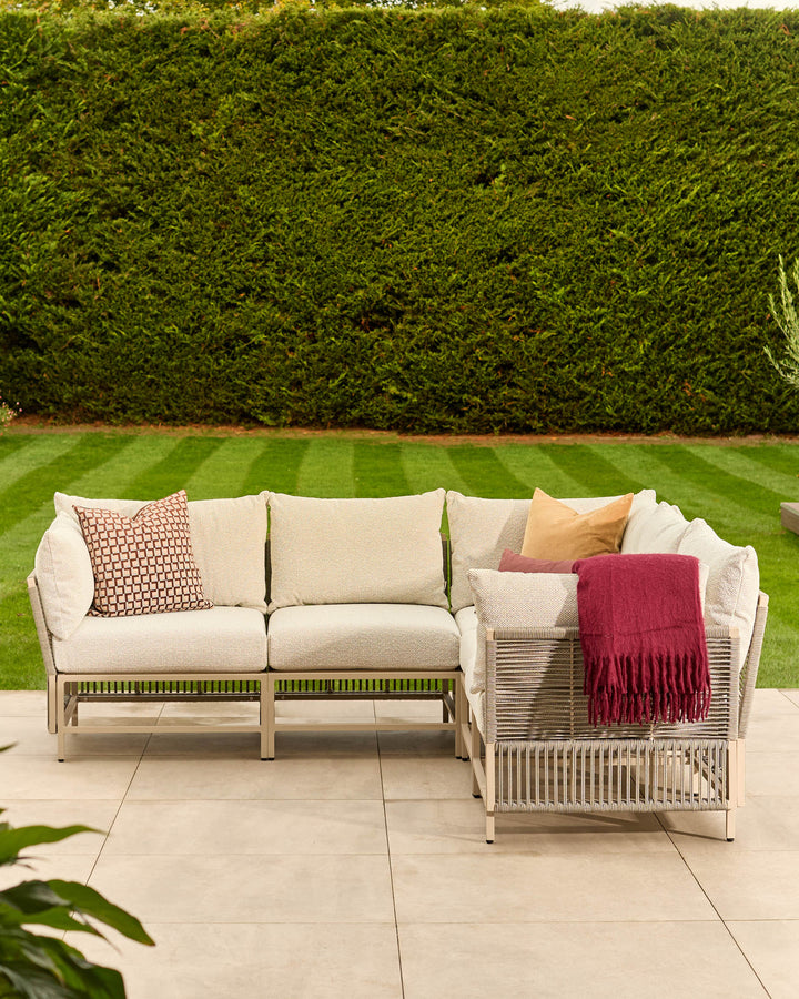 Minori garden large corner bench in natural finish with cream cushions, decorative pillows, and burgundy throw blanket in outdoor patio setting.