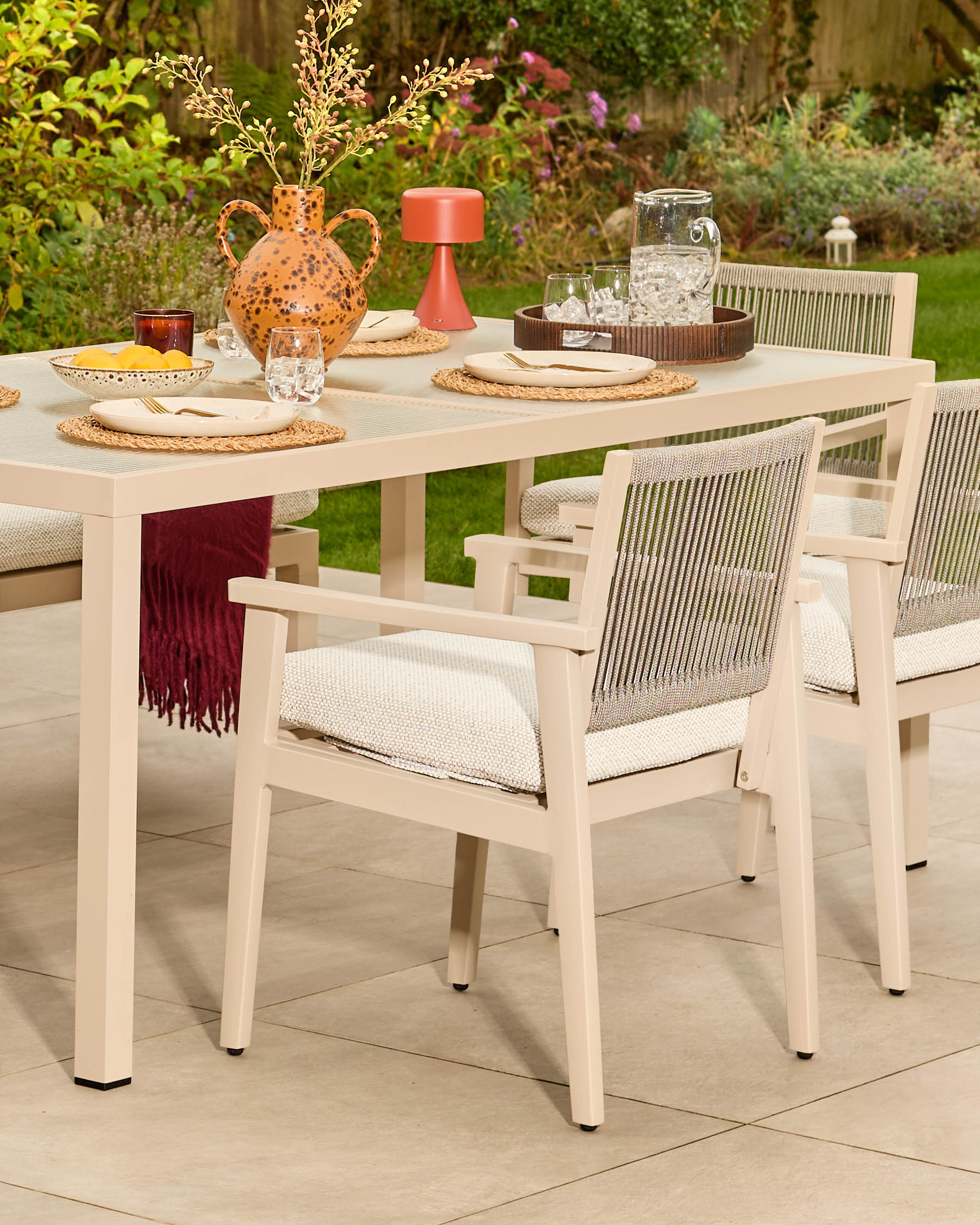 Minori cream garden dining table set for six with woven chairs, styled with tableware and decorative accessories on a patio.