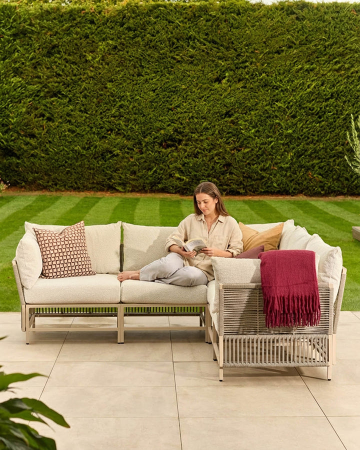Minori natural woven garden corner bench with cream cushions, burgundy throw, and patterned pillows in outdoor setting.