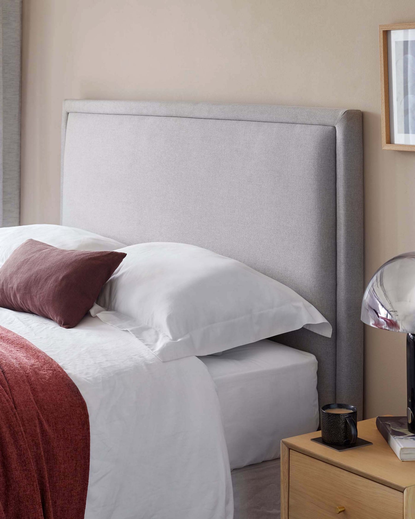 Contemporary styled bedroom featuring a light grey upholstered headboard with clean lines, a natural wood bedside table with a single drawer and a slender metal handle, paired with a modern spherical metal table lamp on top. The bed is dressed with crisp white linens and accented with a terracotta throw and matching pillow. A simple wooden frame with an abstract print hangs on the wall above the table.