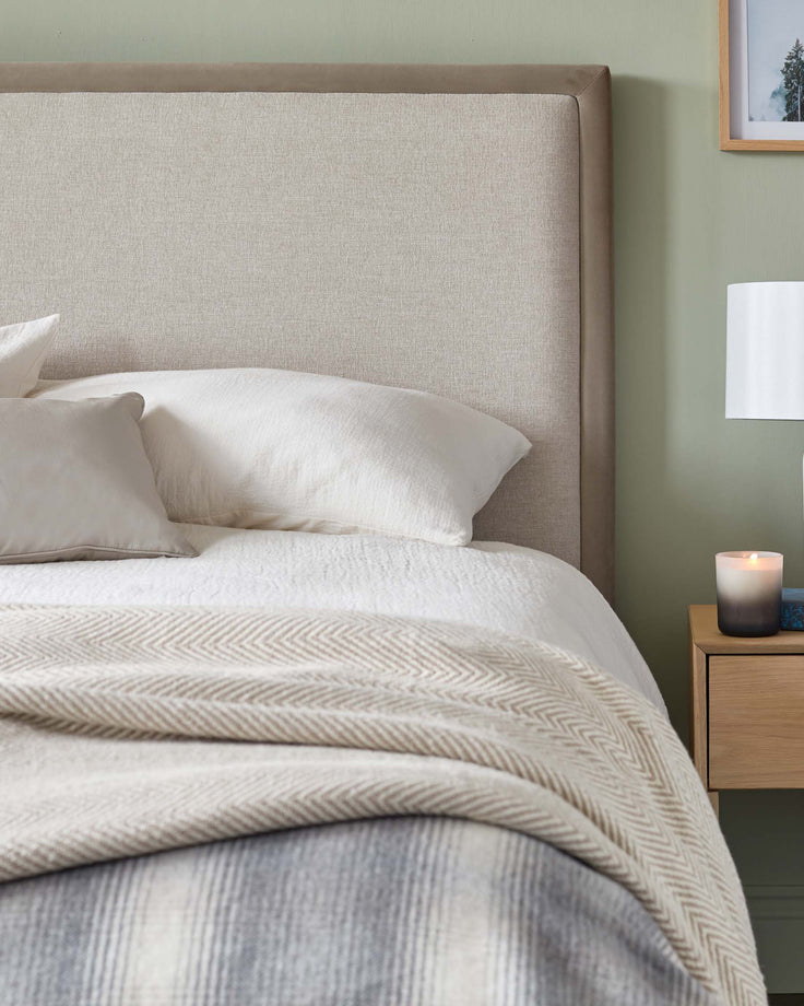 A neutral bedroom with a linen headboard, crisp white bedding and a textured throw. A wooden bedside table features.