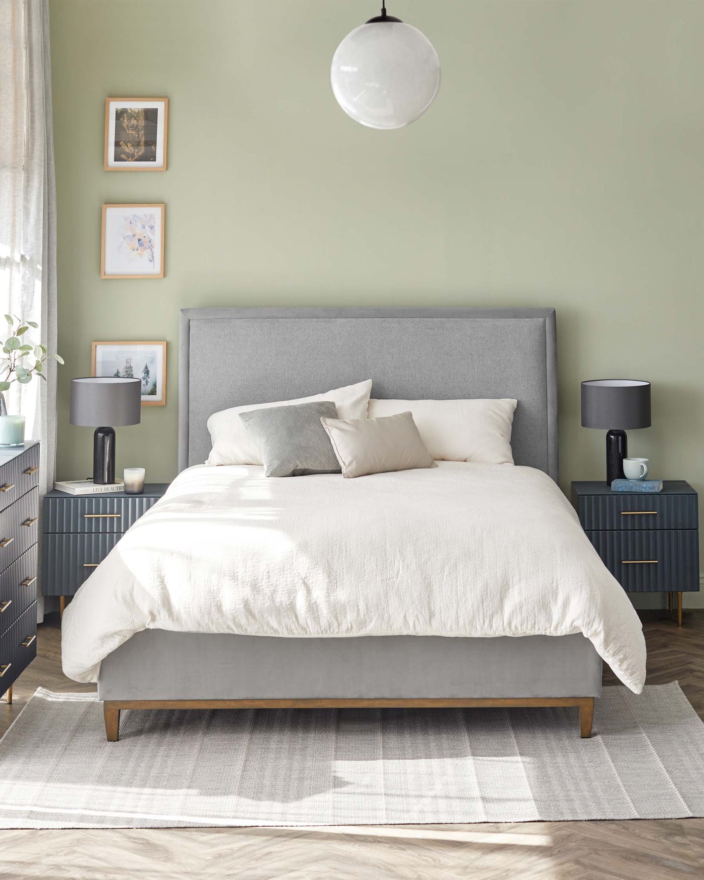 A modern bedroom featuring a grey upholstered bed frame with a high headboard, two dark grey bedside tables with brass handles, and a white area rug underneath the bed. The scene is complemented by minimalist decor, including framed wall art, a white globe pendant light, and simple bedding in neutral tones.