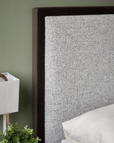 Upholstered headboard in grey fabric with a dark brown trim, on a bed with white bedding, with a lamp on the side table.