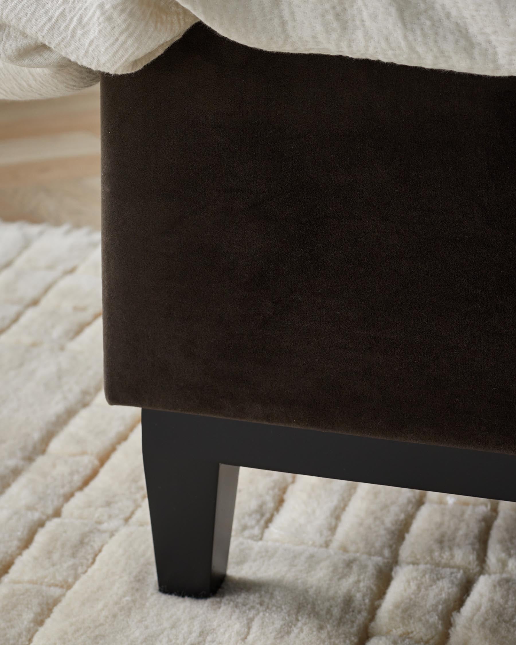 Close-up of a dark brown upholstered bed base with black legs on a cream rug.