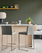 Two grey bar stools with metal legs at a kitchen island with a marble countertop and green walls.