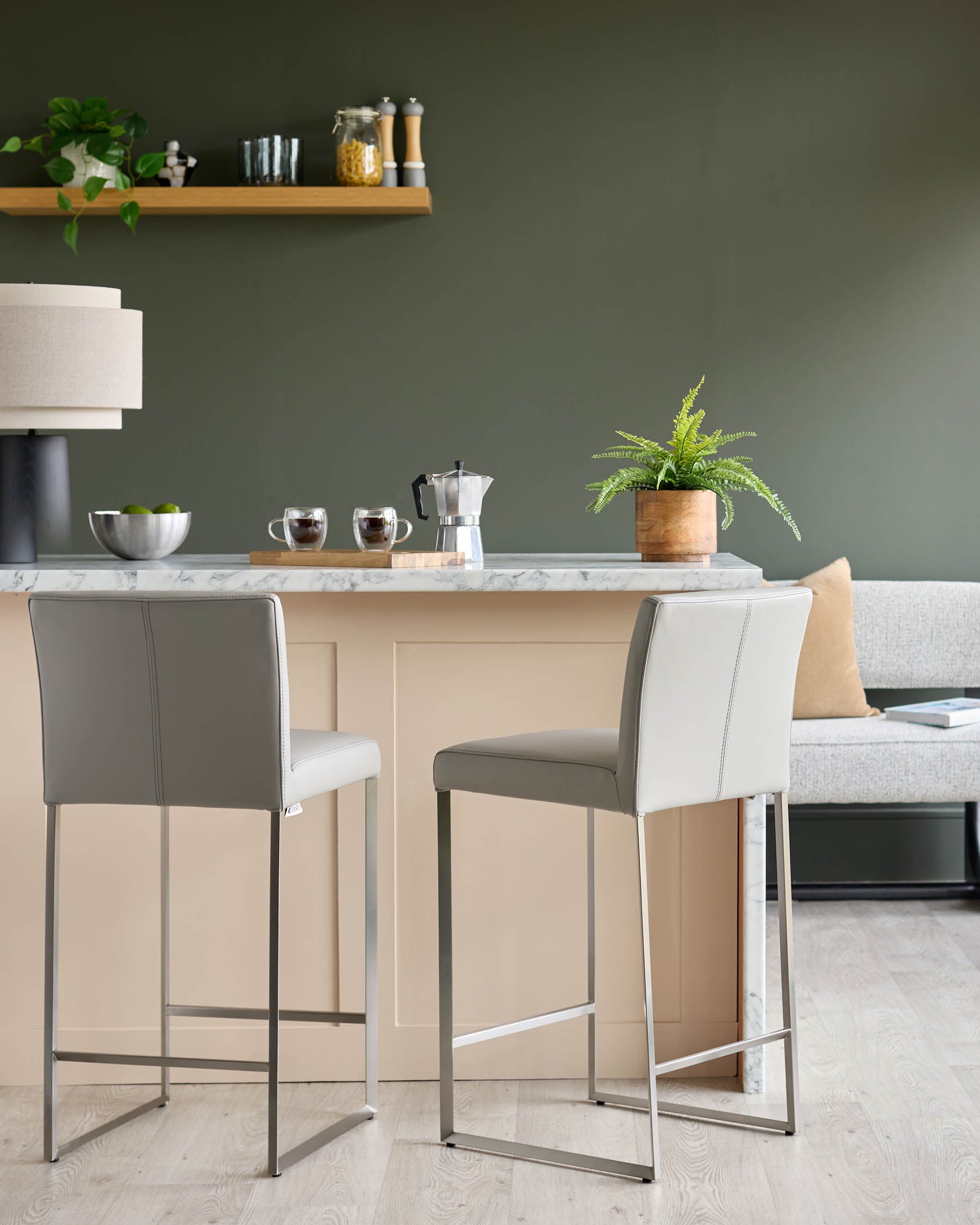 A kitchen island with two grey bar stools. Coffee cups, a plant, and coffee maker sit on the marble top.