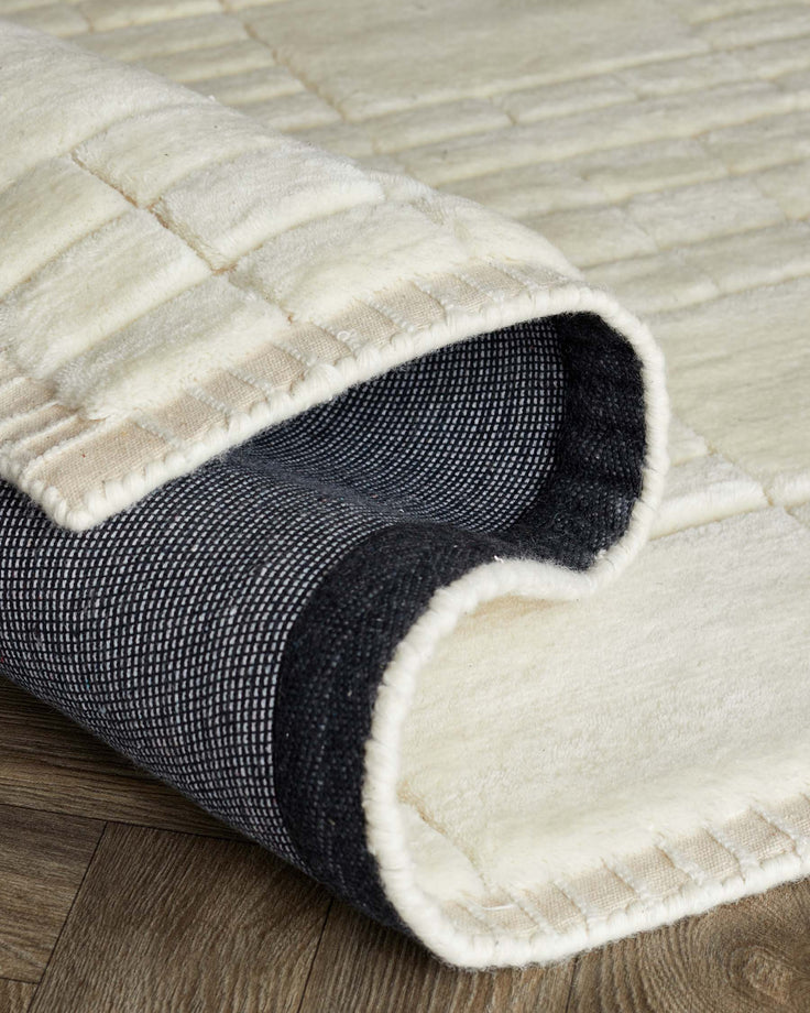 Plush white area rug with subtle geometric textures, partially rolled to reveal a black non-slip backing, presented on a wooden floor.