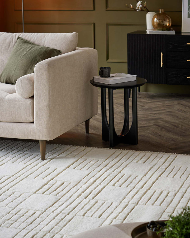 A contemporary beige upholstered armchair with plush cushioning and a green accent pillow rests on a textured cream area rug with geometric patterns. Beside it, a minimalist round black side table with a thin metal frame holds a coffee mug and a small stack of books, complementing a sleek black cabinet with golden handles in the background.