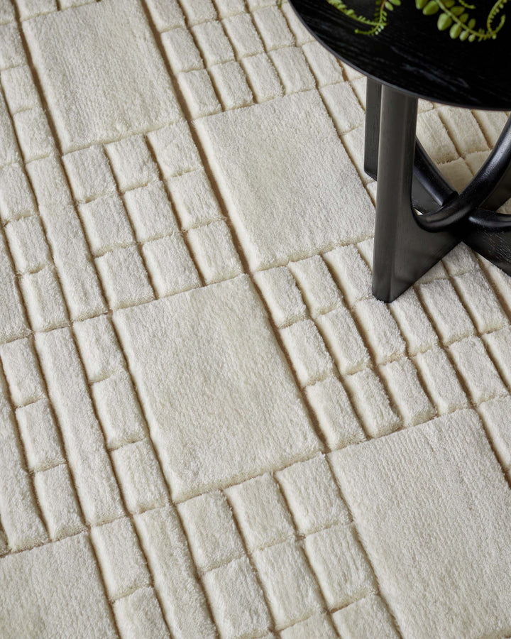 The image shows a textured cream-colored area rug with a geometric pattern of raised squares and rectangles. A small portion of a sleek, round black table with a reflective surface and unconventional Y-shaped legs can be seen, suggesting a modern and minimalistic style.