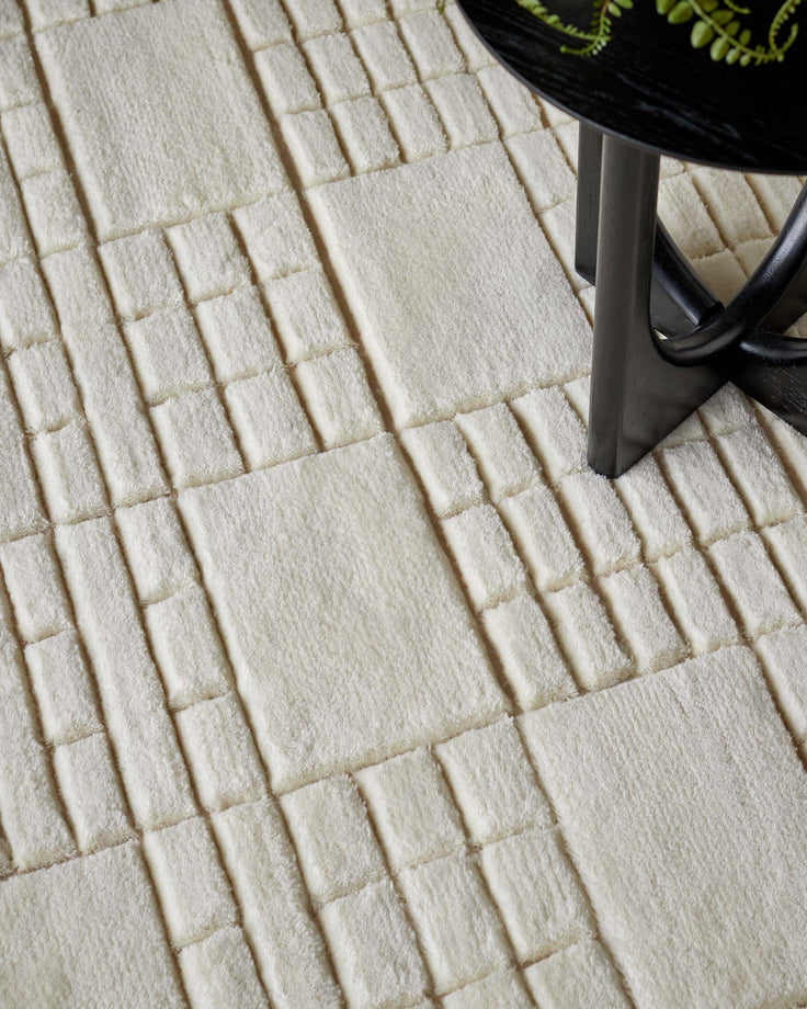 The image shows a textured cream-colored area rug with a geometric pattern of raised squares and rectangles. A small portion of a sleek, round black table with a reflective surface and unconventional Y-shaped legs can be seen, suggesting a modern and minimalistic style.