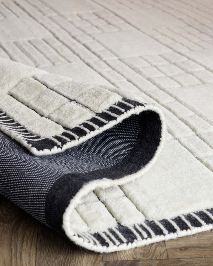 Cream and charcoal patterned area rug with a geometric design, partially rolled up to show texture and thickness, on a wooden floor.