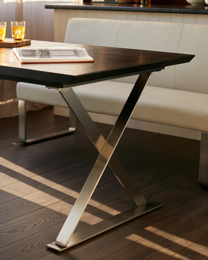 A modern dark wood dining table with striking stainless steel crossed legs, paired with a stylish cream leather bench seat.