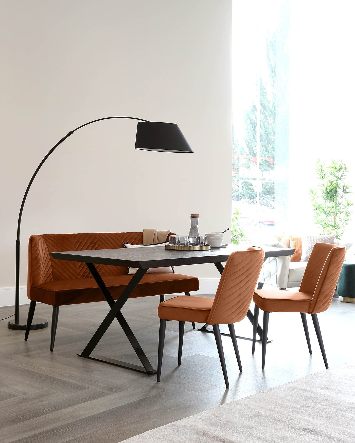 Modern dining room with a rectangular black table with X-shaped metal legs, four upholstered chairs in terracotta with black metal legs, and an elegant, curved black floor lamp with a large shade leaning over the table.