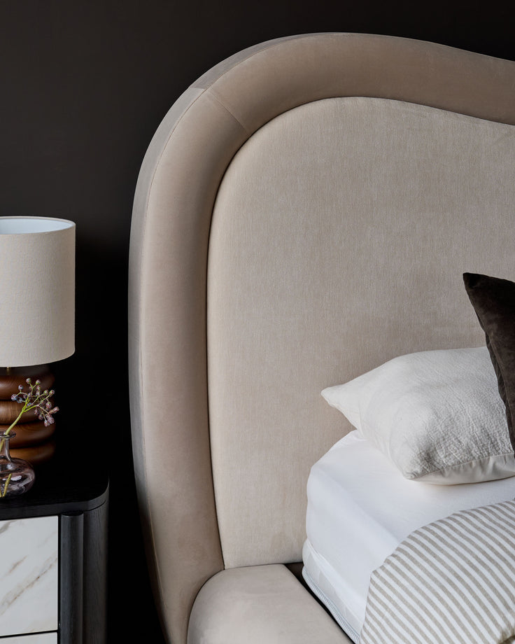 Naomi king-size ottoman bed in champagne velvet with curved headboard and white bedding, styled with bedside lamp.