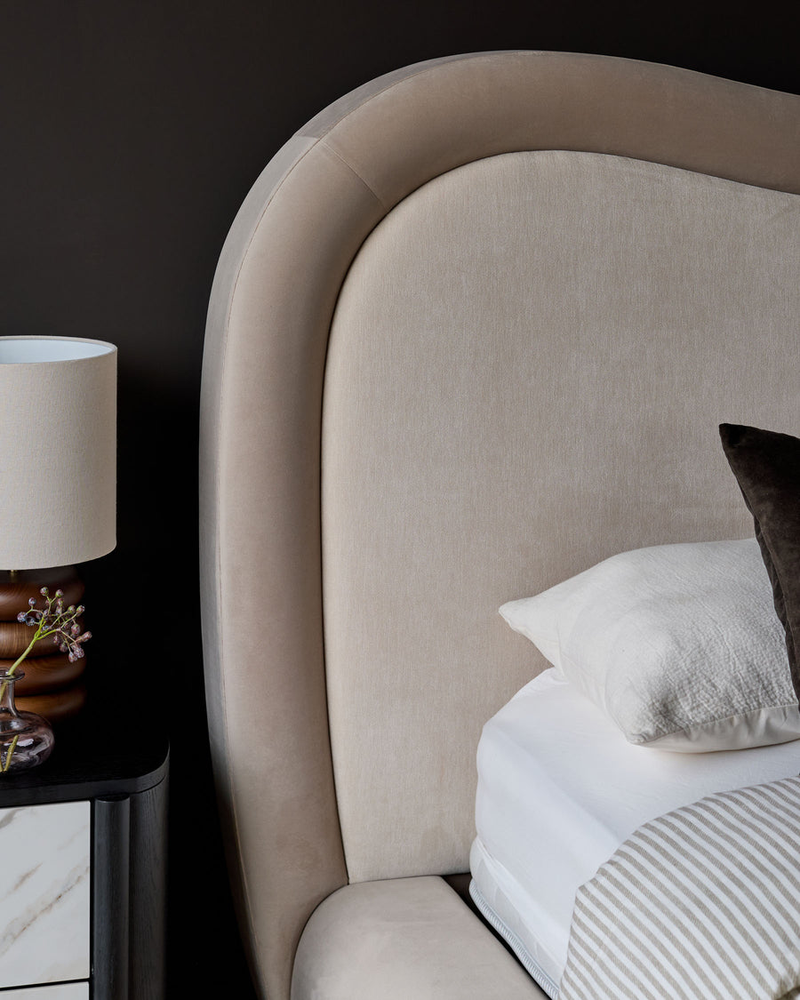 Naomi king-size ottoman bed in champagne velvet with curved headboard and white bedding, styled with bedside lamp.